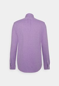 Košile s dlouhým rukávem ve světle fialové barvě, vyrobená z měkké tkaniny. Má manžetu na knoflík a zadní záhyb pro větší volnost pohybu.