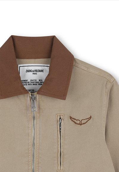 Giacca beige con colletto marrone, cerniera anteriore argentata, tasca verticale con cerniera e dettaglio ricamato a forma di ala marrone sul petto.