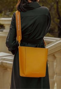 Żółta skórzana torba typu bucket o gładkiej fakturze i regulowanym pasku, z metalowym zapięciem i wytłoczonym logo marki.