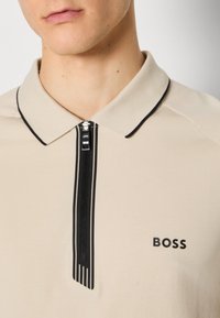 Polo beige avec des bordures noires et fermeture éclair. Présente le logo "BOSS" sur le côté gauche. Le tissu a une texture lisse.