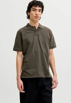 Jaunas vyras stovi, dėvėdamas tamsiai alyvuogių žalios spalvos trumpomis rankovėmis polo marškinėlį ir juodas kelnes, priešais paprastą šviesią foną.