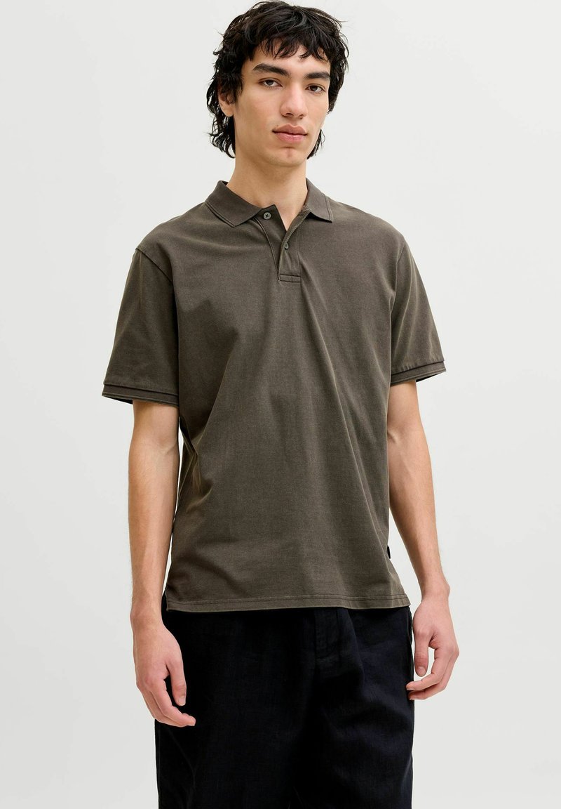 Jeune homme debout portant un polo à manches courtes vert olive foncé et un pantalon noir, sur un fond clair uni.