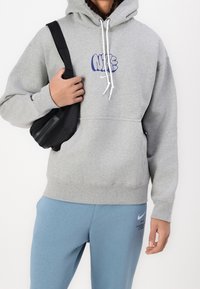 Šedá mikina s kapucí a modrým logem Nike, šňůrky a klokaní kapsa, v kombinaci se světle modrými tepláky s logem Nike. Černá taška.
