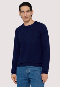 Marinblå sweatshirt med rund hals, ribbade ärmslut och fåll, mjuk textur, kombinerad med ljusblå denimjeans. Diskret logotyp på bröstet.