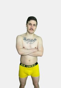 Boxer brief gialli con un elastico nero etichettato "MR UKKO." Modello con tatuaggi e senza camicia, braccia incrociate, su uno sfondo bianco.