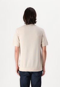 Beige kortermet polo med teksturert strikk, rett hem og subtil krage. Båret med mørkeblå jeans, sett fra ryggen.