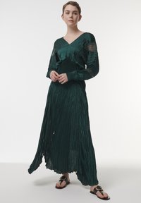 Largo vestido plisado en verde profundo, con escote en V, mangas largas y acentos de encaje transparente en las mangas. Se lleva con sandalias planas.