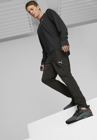 T-shirt à manches longues noir avec le logo Puma gris, associé à un pantalon noir arborant un logo blanc. Baskets grises avec accents rouges, surfaces texturées.