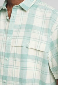 Camicia a maniche corte a quadretti in verde menta chiaro e crema. Presenta una chiusura con bottoni, due tasche sul petto e un tessuto morbido e leggero.