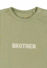 Šviesiai žalia vaikiška marškinėliai, pagaminti iš tekstūruotos medžiagos, su apvaliu iškirpimu ir žodžiu "BROTHER" ryškiai išsiuvinėtu balta spalva.
