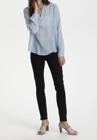 Frau, die eine hellblaue gemusterte Langarmbluse, schwarze enge Hose und beigefarbene offene Absatzsandalen trägt, vor einem schlichten weißen Hintergrund.