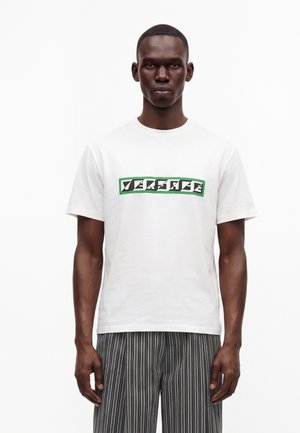 Άνδρας που φοράει λευκό μπλουζάκι Versace με πράσινο και μαύρο λογότυπο και μαύρο-λευκό κάθετα ριγέ παντελόνι, στέκεται μπροστά σε λευκό φόντο.