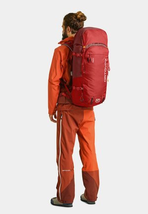 Roter technischer Rucksack mit mehreren Riemen und Taschen, getragen von einer Person in einer orangefarbenen Jacke und Hose, vor einem schlichten Hintergrund.