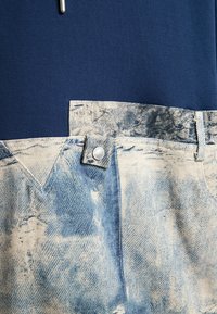 Tela de mezclilla en azul claro con detalles desgastados y deshilachados. Cuenta con una sección superior en azul oscuro, con un botón de metal y acentos en la cinturilla.