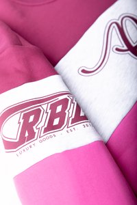 Ružová a biela tričko s výrazným textom "RBL" a "LUXURY GOODS" v bordovej farbe. Bavlnený materiál s hladkou textúrou a kontrastnými farbami.