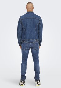 Giacca in denim e jeans blu medio con vestibilità classica, dotati di tasche posteriori e accessori in metallo. Indossati con sneakers bianche e nere.