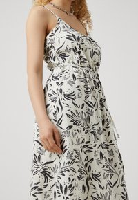 Robe blanche à motif de feuilles noires, bretelles spaghetti, taille ajustée et jupe fluide. Fabriquée en tissu léger à texture lisse.