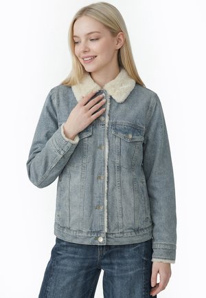 Ung kvinde iført en blå denimjakke med hvidt fleecekrave og -manchetter, smilende og kigger ned med sin højre hånd nær brystet.