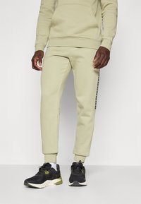 Haut à capuche en coton vert clair avec une poche à l'avant, associé à un jogging assorti avec une bande latérale. Des baskets noires et olive complètent le look.