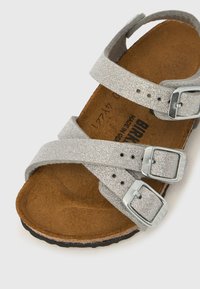 Hõbedase sädelevate sussidega, millel on kolm reguleeritavat pandlaga rihma ja pruun kujundatud tallaga, valgel taustal.