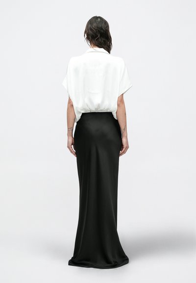 Žena s tamnom kosom do ramena koja nosi bijelu bluzu kratkih rukava uvučenu u dugu crnu suknju, stoji okrenuta leđima.