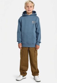 Felpa con cappuccio in cotone blu con tasca frontale e logo, abbinata a pantaloni cargo marroni a taglio ampio e sneaker bianche con dettagli verdi.