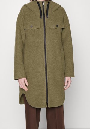 Oliwkowozielony wełniany płaszcz z kapturem, zapinany na zamek z przodu, z kieszeniami na patki zapinanymi na guziki oraz bocznymi rozcięciami, noszony na brązowych spodniach.