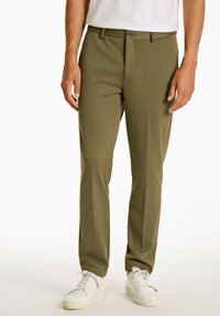 Olivgrüne, figurbetonte Hose mit Reißverschluss, einem Knopfverschluss und seitlichen Taschen. Getragen mit weißen Sneakers vor einem neutralen Hintergrund.