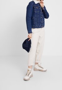 Person med en blå denimjacka med vit sherpakrage, krämfärgade byxor, vita vandringskängor, som håller en blå denimbucket hat mot en vit bakgrund.