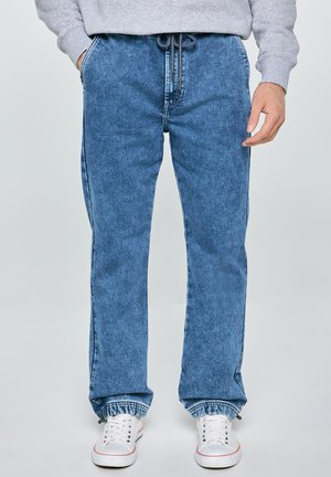 Muž v modrých džínových jogger kalhotách s elastickými manžetami a bílými teniskami, ruce v kapse, stojící na jednobarevném pozadí.