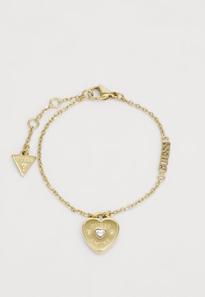 Złota bransoletka z łańcuchowym wzorem, z sercowym wisiorkiem wyrzeźbionym napisem "GUESS AMORE" oraz małym trójkątnym charmsami.