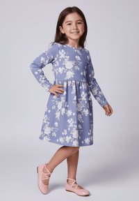 Jasnoniebieska sukienka z długim rękawem, z białymi kwiatowymi wzorami, dopasowaną górą i rozkloszowanym dołem. Zestawiona z jasnoróżowymi butami na krzyż.