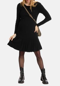 Schwarzes Strickkleid mit langen Ärmeln und plissiertem Saum, kombiniert mit schwarzen Knöchelstiefeln und einer kleinen Umhängetasche.
