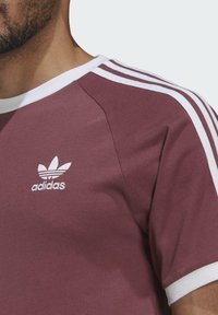 Burgundijas kokvilnas T-krekls ar baltu apkakli un trim baltām svītrām uz piedurknēm. Priekšpusē ir izšūta balta logo. Īso piedurkņu dizains.