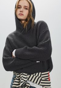 Túlméretezett, sötétszürke kötött kapucnis pulóver texturált mintával. Tágas kapucnival és bordázott mandzsettával rendelkezik. Fehér ing fölött viselve, csíkos nyakkendővel párosítva.