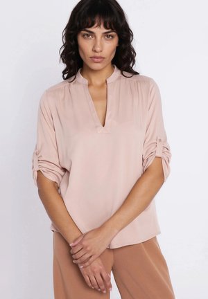 Kobieta o falowanych ciemnych włosach, ubrana w jasnoróżową bluzkę z podwiniętymi rękawami i beżowe spodnie, stoi z rękami skrzyżowanymi przed sobą.