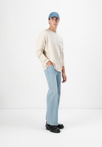Sweatshirt beige avec logo, jeans bleu clair et chaussures à plateforme noires. Le modèle porte une casquette ajustée bleu clair. Tenue simple et décontractée.
