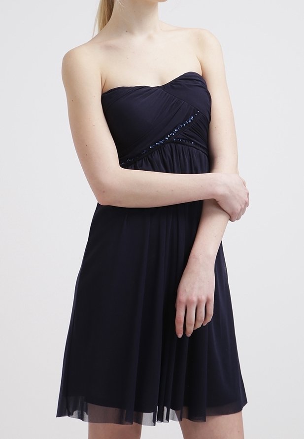 Femme portant une robe noire sans manches et sans bretelles avec un corsage froncé et un subtil détail de sequins au niveau de la poitrine, debout, les bras croisés.
