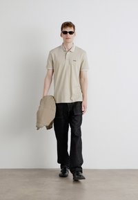 Beige Poloshirt mit Kragen, schwarze Cargo-Hose und Sonnenbrille. Das Model hält eine beige Jacke und steht vor einem neutralen Hintergrund.