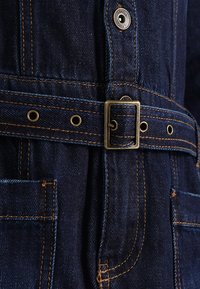Jeansoverall i mörkblå färg, med enkel design, framträdande sömmar, sidofickor och ett bälte med metallspänne.