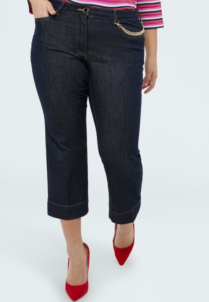 Kobieta w krótkich, ciemnych dżinsach z detalem złotego łańcuszka i czerwonych szpilkach, ręka oparta w kieszeni.