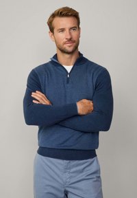 Maglione con zip di colore blu navy, in maglia testurizzata, con colletto alto, maniche lunghe e polsini e orlo a coste. Indossato sopra una camicia bianca.