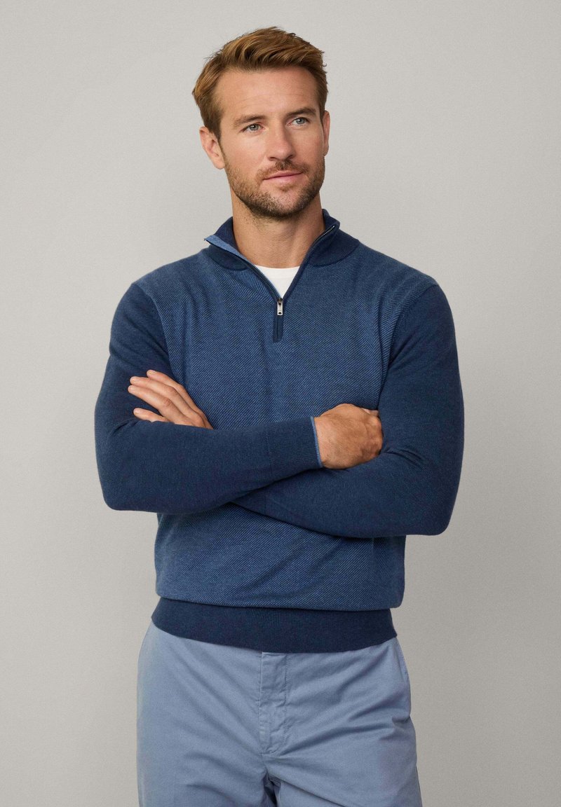Maglione con zip di colore blu navy, in maglia testurizzata, con colletto alto, maniche lunghe e polsini e orlo a coste. Indossato sopra una camicia bianca.