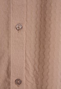 Camicia in tessuto beige con un motivo a zigzag testurizzato, completata da un dettaglio con bottone rotondo e accenti cuciti lungo la patta.