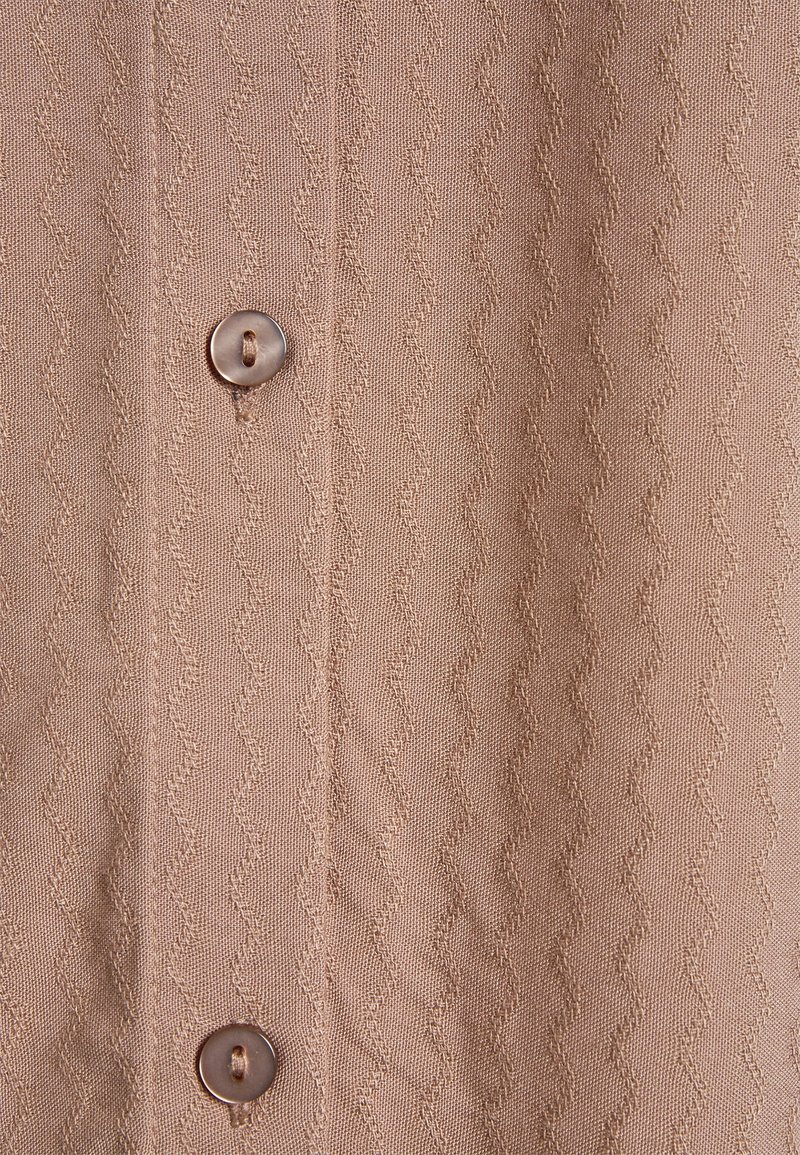 Camicia in tessuto beige con un motivo a zigzag testurizzato, completata da un dettaglio con bottone rotondo e accenti cuciti lungo la patta.