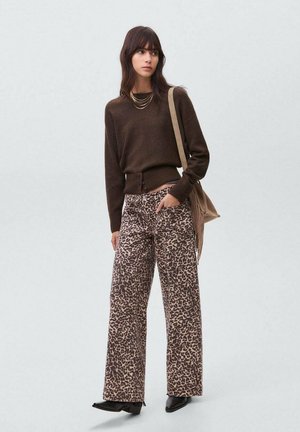 Mujer con cabello castaño oscuro, vestida con un suéter marrón, pantalones anchos de estampado de leopardo, zapatos negros y un bolso de hombro beige, de pie contra un fondo liso.