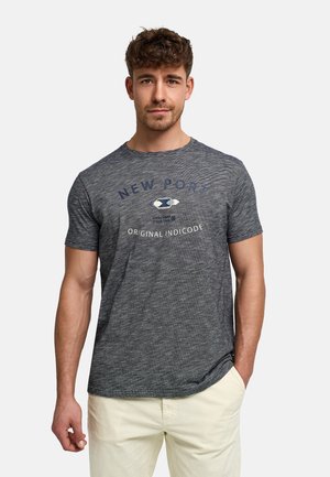 Uomo con capelli corti e ricci che indossa una maglietta grigia con la scritta "New Port Original Indicode" e pantaloni chiari, in piedi con una mano in tasca.