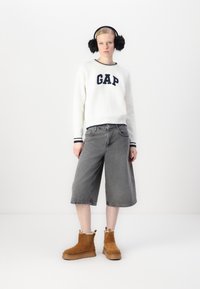 Valge kootud sviiter, millel on "GAP" logo, hallad denimplikaadid ja beežid nubukist pahkluu saapad, mille sees on karvane vooder. Mustad kõrvaklapid täiendavad välimust.