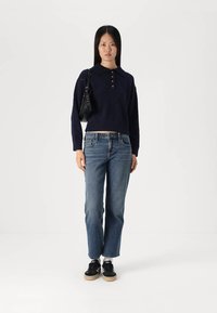 Mornarska pletena pulover s tri gumba i kragnom, u kombinaciji s svijetloplavim trapericama i crnim tenisicama. Model drži malu crnu torbicu.