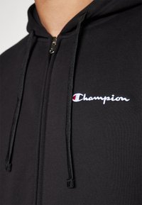 Schwarze Zip-Up-Hoodie aus Stoff mit glatter Textur; verfügt über ein gesticktes 'Champion'-Logo in Weiß und Rot auf der Brust.